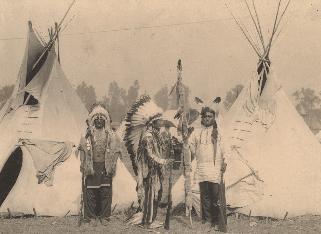 Une photographie vintage en noir et blanc montre aux Amérindiens en robe traditionnelle debout devant les tipis, représentant le patrimoine culturel central du débat de dénomination de l'équipe.